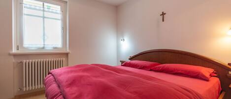 A bedroom with a bed, a window, a radiator, and a cross on the wall.
