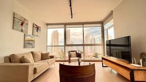 A modern living room with a sofa, a coffee table, and a TV. Large windows offer a view of tall buildings.
