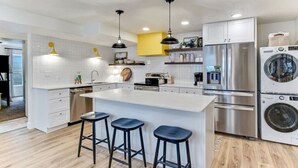 Una cocina moderna con armarios blancos, electrodomésticos de acero inoxidable, una isla central con taburetes y luminarias colgantes.