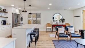 Una cocina moderna con armarios blancos, una isla central con taburetes, una zona de comedor con mesa redonda y sillas, y un espejo grande.