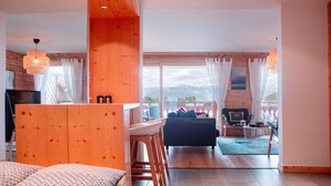A spacious living area with a wooden interior, a dining table, and a sofa. A large window offers a view of mountains.