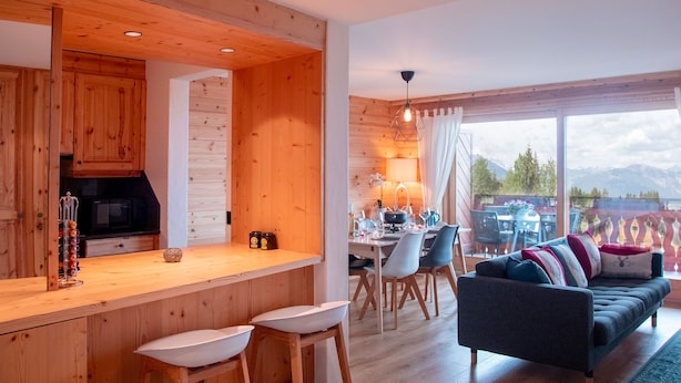 A modern kitchen with wooden accents, a dining area with a table and chairs, and a sofa in a living room with a view of mountains.