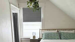 A bedroom with a bed, a bedside table, a lamp, a window with curtains, and a hanging plant.