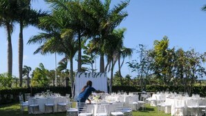Montaje para un evento al aire libre con mesas redondas cubiertas con manteles blancos y sillas, rodeado de palmeras.