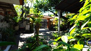 Un patio con techo de baldosas, rodeado de abundante vegetación y un sendero que lleva a un edificio con puerta roja.