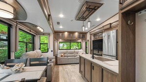 A modern RV interior with a dining area, seating, kitchen, and large windows offering views of greenery. - Luxury Creekside Retreat: RV in Gatlinburg,Tn (Gatlinburg)