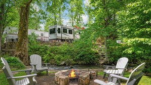 A campsite with a fire pit surrounded by Adirondack chairs, a white RV, and a stone bridge over a stream. - Luxury Creekside Retreat: RV in Gatlinburg,Tn (Gatlinburg)