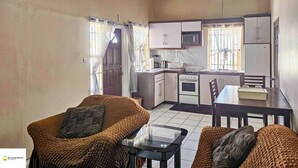 Un espacio habitable compacto con kitchenette, dos sofás de mimbre, una mesa de centro de vidrio y una mesa de comedor.