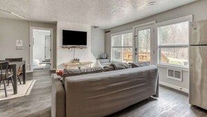 A modern living room with a large gray sofa, a flat-screen TV mounted on the wall, and a dining area with a table and chairs. - Riverbend Cabin Hot Tub, River Front Dog Friendly (Rhododendron)