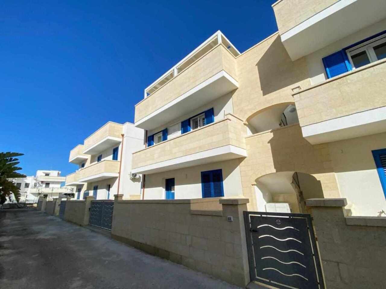 A row of modern, multi-story residential buildings with balconies, a gated entrance, and a clear blue sky.
