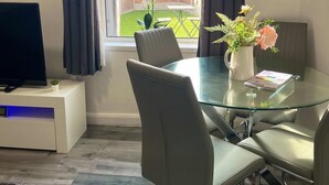 Uma sala de estar moderna com uma mesa de jantar de vidro, cadeiras estofadas cinzas, uma televisão de tela plana em um armário branco e vista para um quintal com cerca de madeira e vegetação.