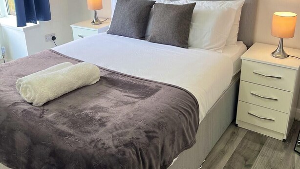 Cama bem arrumada com manta e travesseiros cinzas, duas mesas de cabeceira com luminárias e uma janela com cortinas azuis.