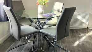 Uma área de jantar moderna com uma mesa de vidro rodeada por cadeiras estofadas cinzentas. Um vaso branco com flores está sobre a mesa, e há uma estante branca com uma planta e um livro ao fundo.