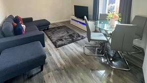 Uma sala de estar com sofá, mesa de vidro, televisão e um canto de jantar com cadeiras.