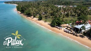 Una vista aerea di una spiaggia con palme, una riva sabbiosa e alcuni edifici con tetti rossi.