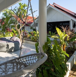 Un fauteuil suspendu dans un jardin, entouré de différentes plantes, avec un bâtiment visible au loin.