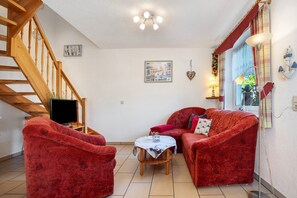 Un salon avec un escalier en bois, un téléviseur à écran plat, des canapés rouges, une petite table et un luminaire au plafond.
