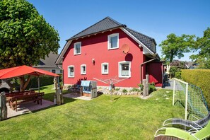 Une maison rouge avec un toit en pente, une antenne parabolique et un jardin agrémenté d’un banc et d’une table.