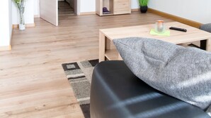 Ein modernes, helles Zimmer mit Holzboden, einer schwarzen Couch, einem kleinen Tisch, einem an der Wand befestigten Fernseher und einer Tür zu einem weiteren Raum.