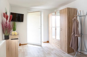 Ein Zimmer mit Holzschrank, Wandfernseher, Kleiderständer mit Bademantel und Topfpflanze mit roten Blättern.