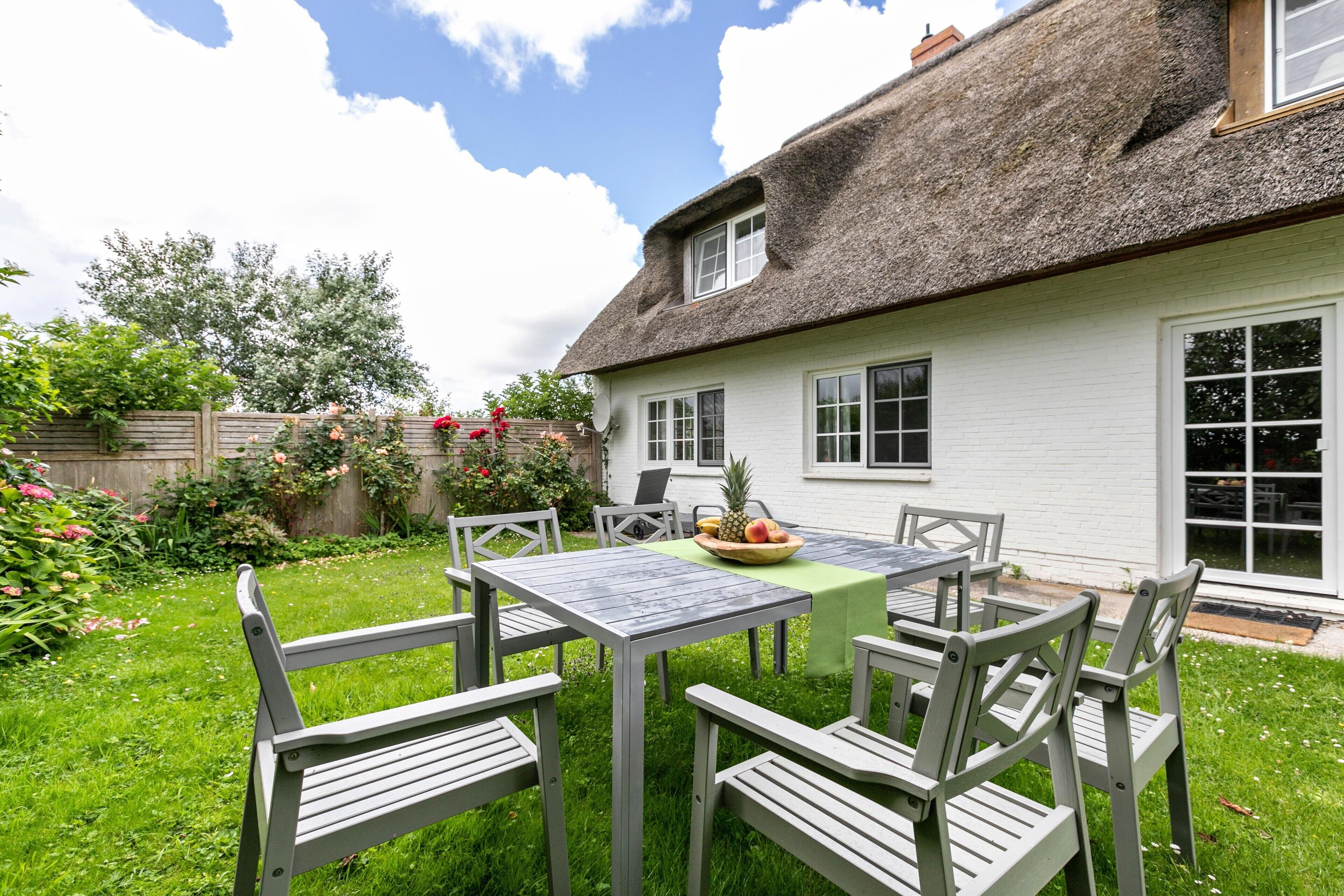 A thatched-roof house with a white exterior, a garden with a table and chairs, and a fruit bowl on the table.