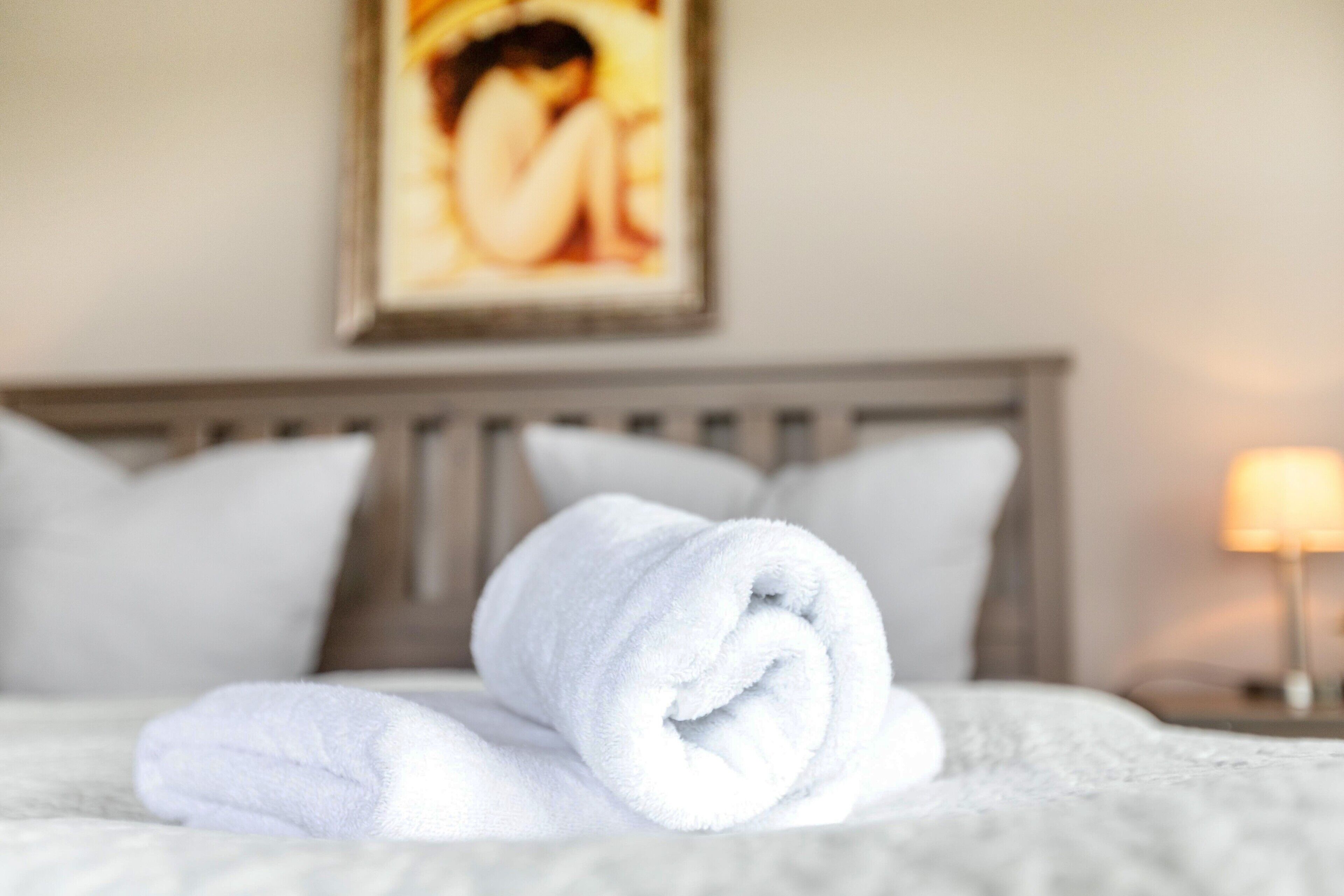 A neatly made bed with a rolled white towel, a wooden headboard, and a framed picture on the wall.