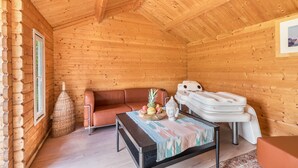 Interior de una cabaña de troncos con un sofá, una mesa con frutero, una estatua de Buda y un sillón de masaje.