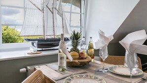 Une table à manger ornée d’un modèle de voilier, d’un panier de fruits, de bouteilles de vin et de verres, prête pour le repas.