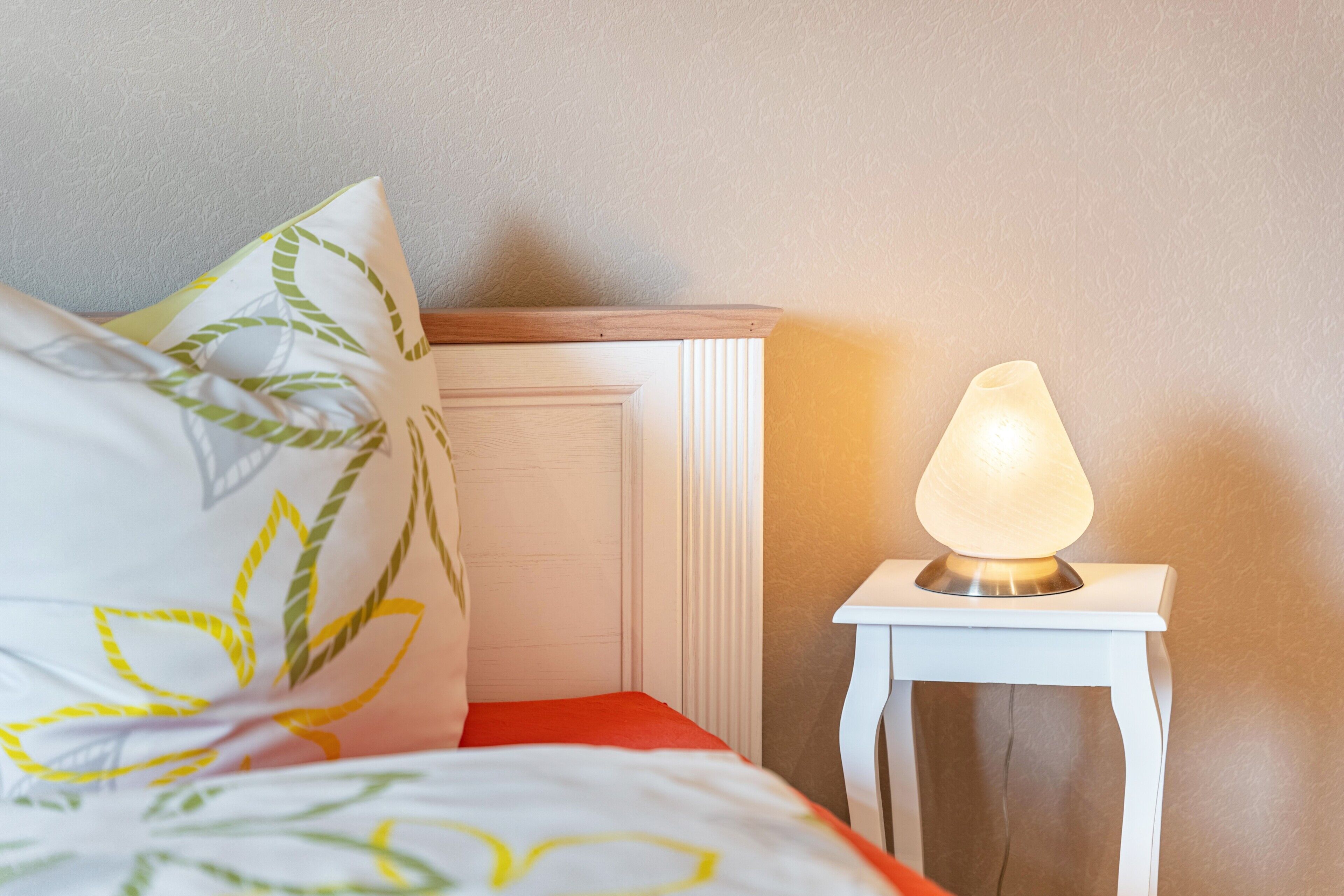 A bed with a white and orange bedspread, a pillow with a leaf pattern, and a bedside table with a lit lamp.