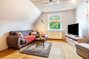 Moderni olohuone, jossa on sohva, sohvapöytä, televisio ja ikkuna, josta avautuu näkymä vehreään maisemaan.
