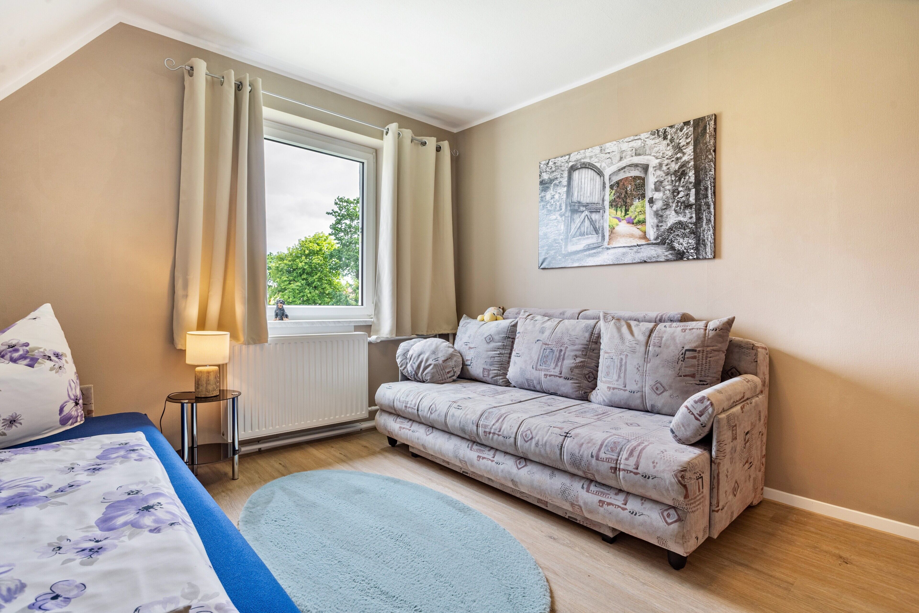 A bedroom with a bed, a sofa, a window with curtains, a radiator, and a painting on the wall.