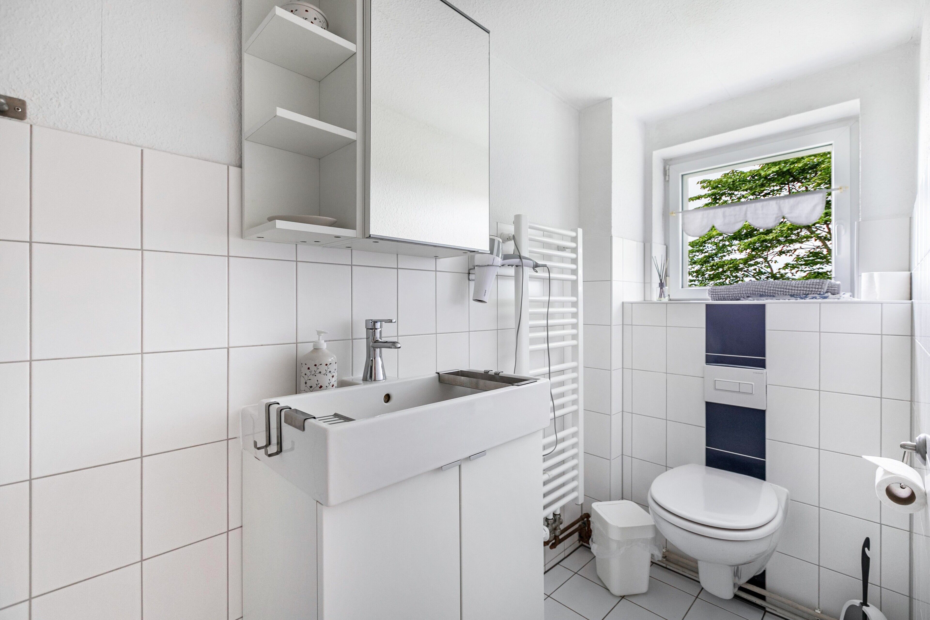 Un baño moderno con un lavamanos blanco, un inodoro, una ventana con vistas a la vegetación y un perchero para toallas.