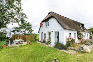 Une maison blanche avec un toit de chaume, un jardin avec des fleurs et des plantes, et une clôture en bois.