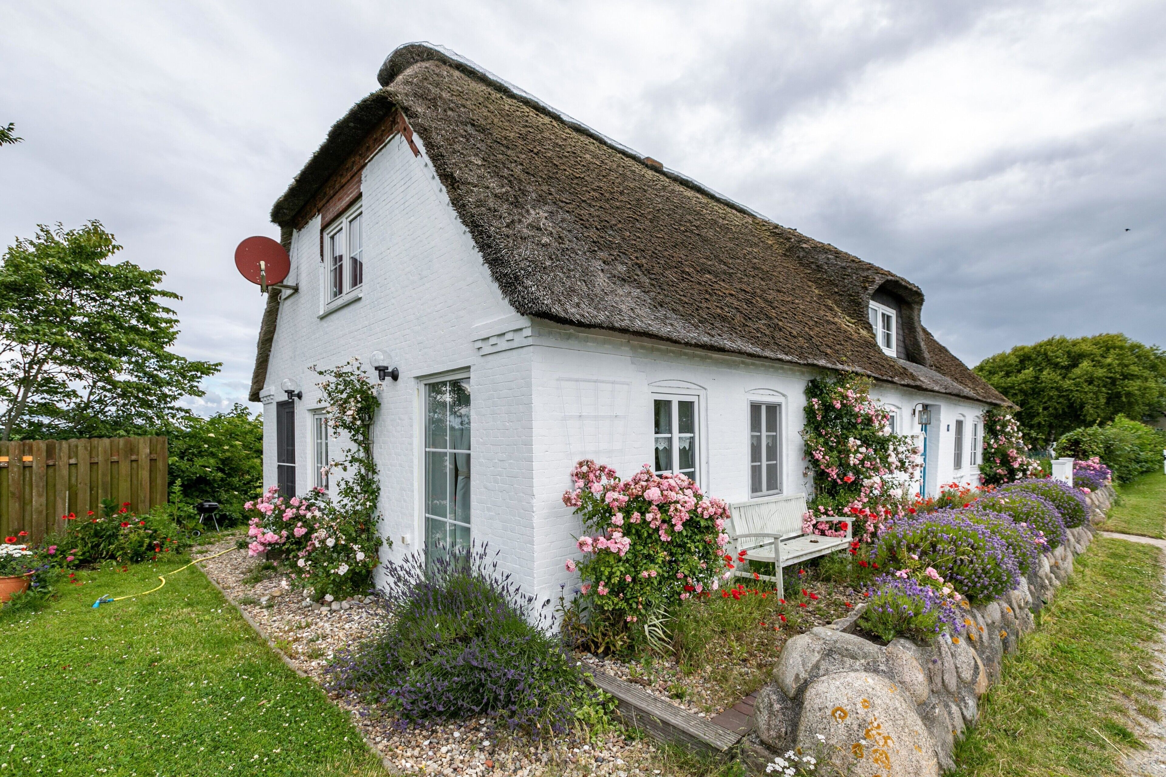 Une maison blanche avec un toit de chaume, entourée d’un jardin où poussent différentes plantes et fleurs.