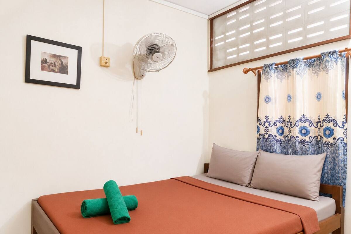 Ein Schlafzimmer mit einem Bett, einem Wandventilator, einem Fenster mit gemusterten Vorhängen und einem gerahmten Bild an der Wand.