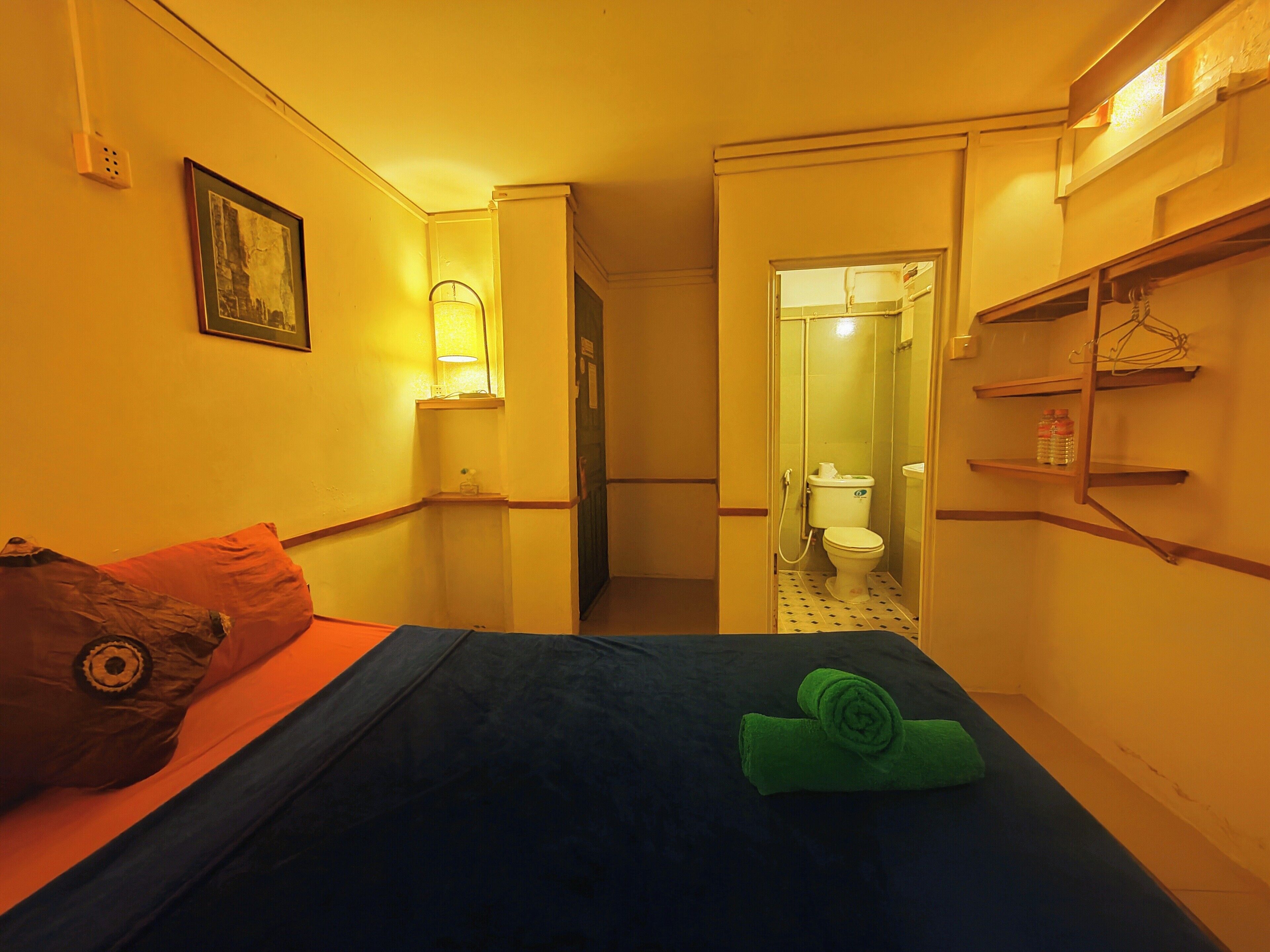 Habitación de hotel estrecha con una cama, una mesita de noche pequeña, un estante en la pared y un baño visible a través de una puerta abierta.