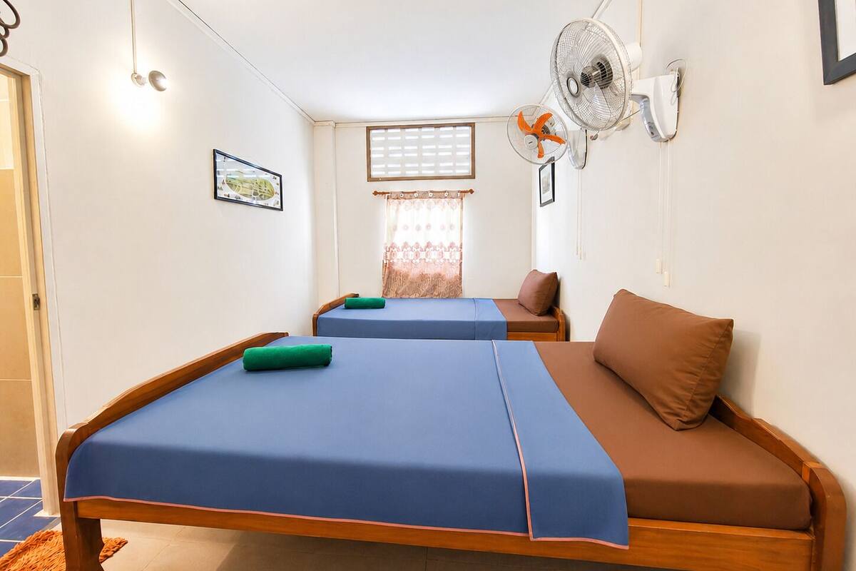 Ein Zimmer mit zwei Betten, einem Vorhangfenster, einem Wandventilator und einem Bilderrahmen.