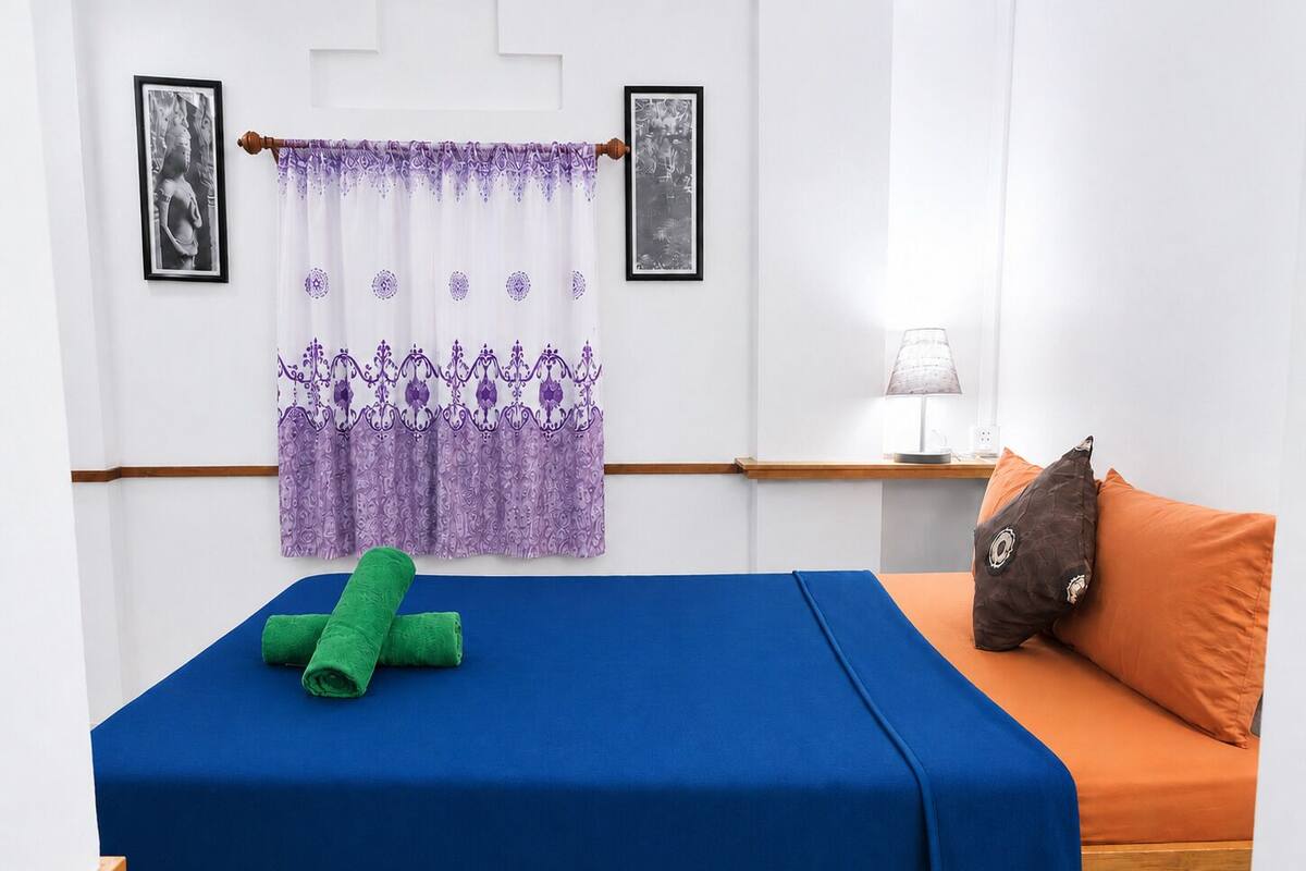 Ein Einzelbett mit blauer Matratze, einem grünen Handtuch, einem orangefarbenen Kissen, einer Lampe auf einem Nachttisch und zwei gerahmten Bildern an der Wand.