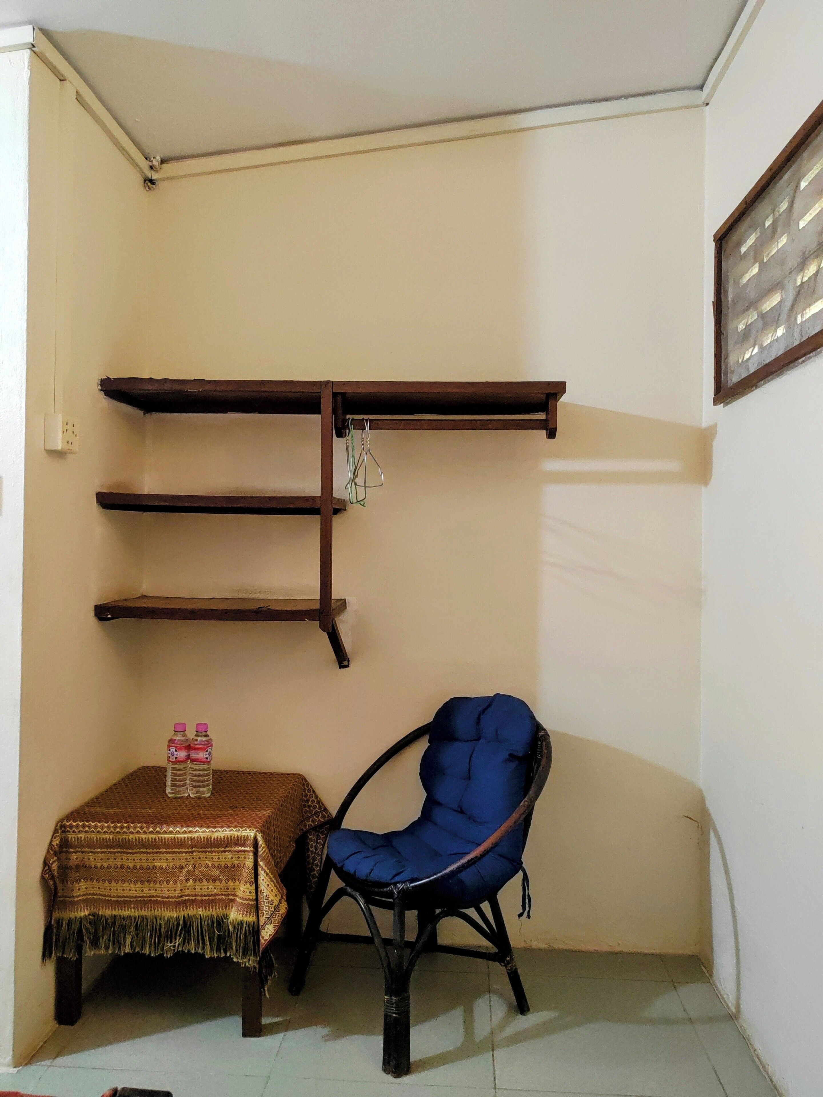 A corner room with a blue cushioned chair, a small table with a patterned tablecloth, and wooden shelves.