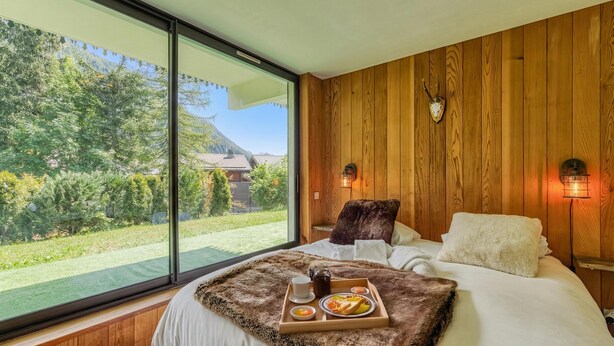 Une chambre avec une grande fenêtre, des murs en bois, un lit avec du linge de lit blanc, un plaid en fourrure et un plateau de petit-déjeuner.