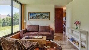 Un salon avec un canapé en cuir marron, une table basse en bois, un tapis en peau de vache, un tableau représentant une montagne encadré, et une console blanche ornée de livres et d’un vase.