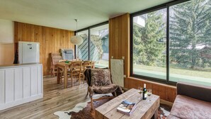 Un intérieur moderne avec un plancher en bois, un coin repas comprenant des chaises et une table, un canapé, un meuble blanc et de grandes fenêtres donnant sur la forêt.