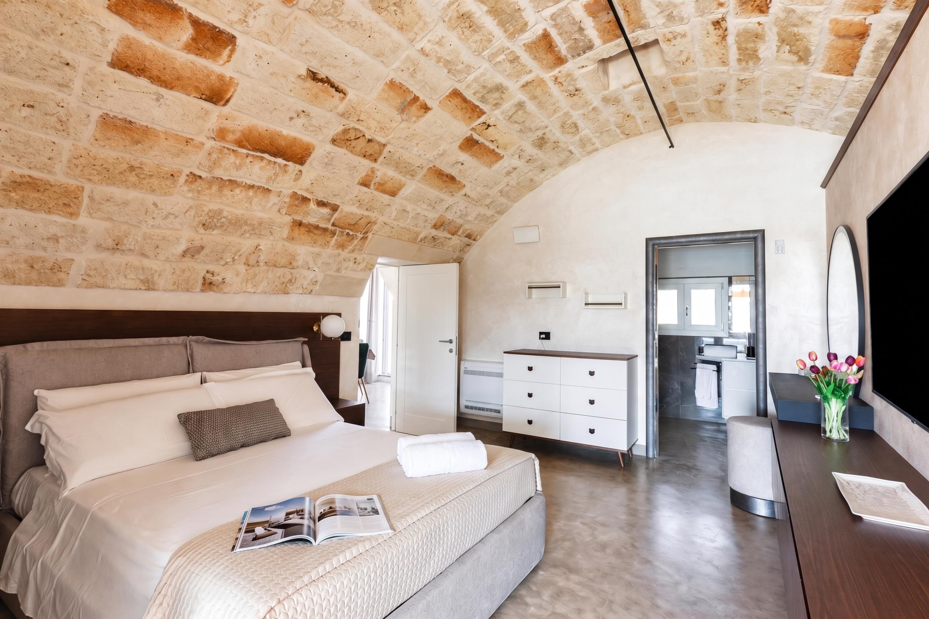 A bedroom with a brick ceiling, a bed with white linens, a bedside table with a magazine, and a view into the bathroom.