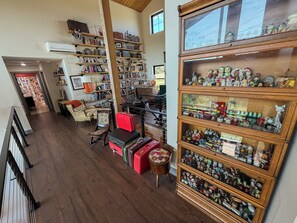 Een ruime kamer met een hoge plafond, houten vloer en een grote collectie boeken en decoratieve objecten op planken. Er staan meerdere stoelen en een klein tafeltje met een rode koffer.