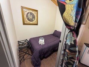 Une petite chambre à coucher avec un lit, une chaise, un tableau encadré au mur et un rideau orné d’un motif de feuilles.