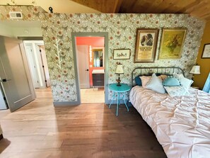Un dormitorio con cama, mesita de noche, cómoda y espejo. Hay una puerta que da a otra habitación.