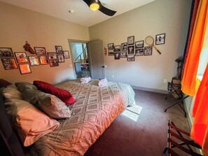 A bedroom with a bed, pillows, a ceiling fan, a wall with framed photos, and a door leading to another room.