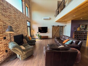 A spacious living room with a brick wall, a flat-screen TV, leather sofas, and a wooden ceiling.