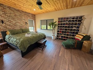 Een slaapkamer met een bakstenen muur, een houten plafond, een bed met groene beddengoed, een houten bankje, een plant en een muur vol hoeden.