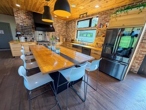 Una cocina moderna con una mesa de madera, sillas blancas, un refrigerador de acero inoxidable y una pared de ladrillo.